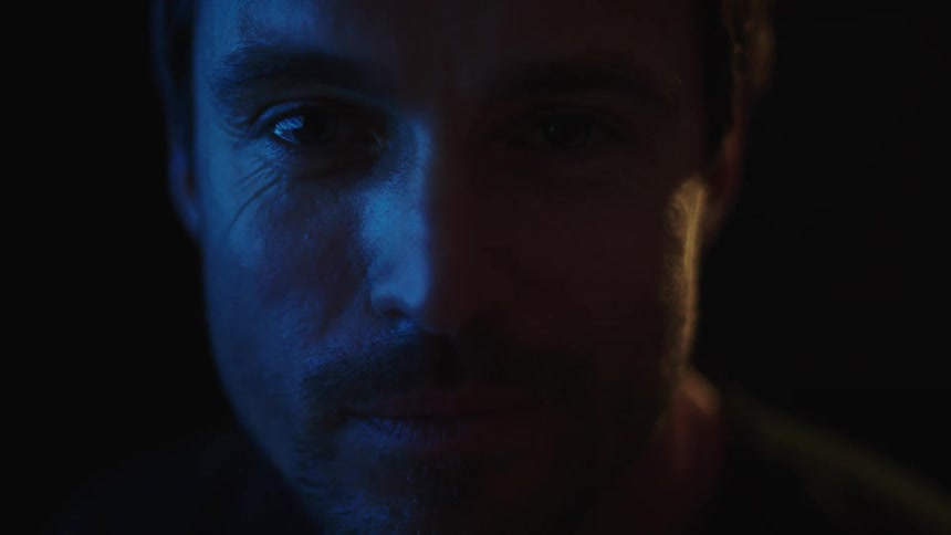 A closeup of a mans face in the dark with a subtle smile as his expression changes and his face is lit by blue and white light