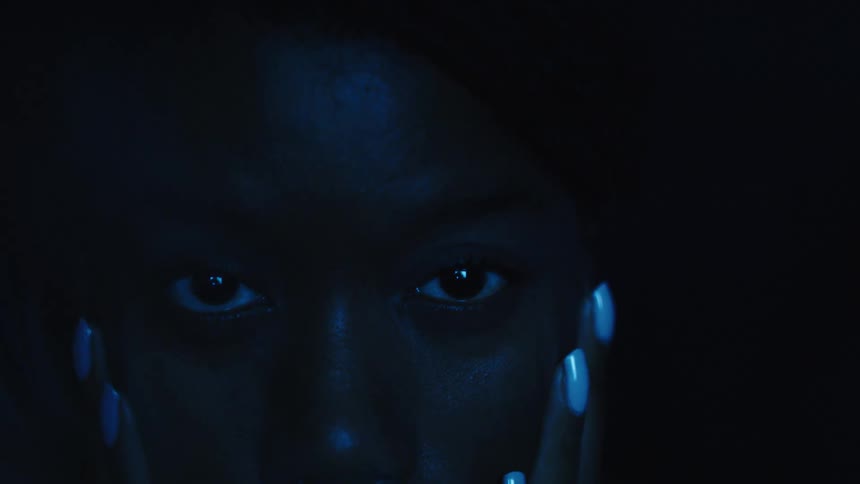 A persons face is seen in a dark room the face is illuminated with a bright blue light and has a serious expression