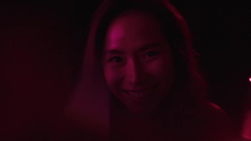 A woman with dark hair is smiling while being lit by a pink and red light