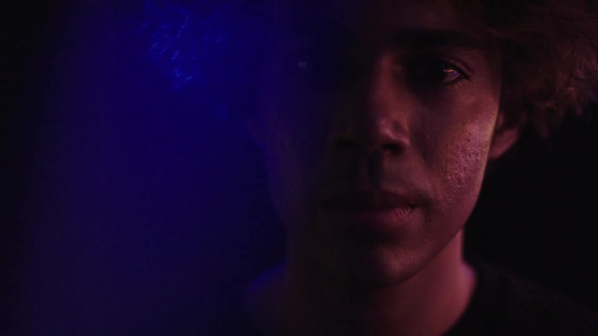 A closeup shot of a mans face is halfhidden in shadow with his features illuminated by a colorchanging light as he looks upwards