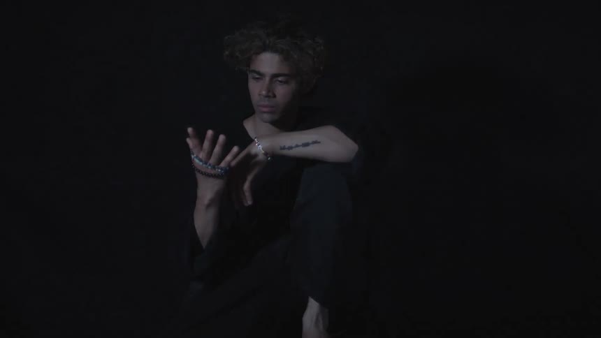 A young man with curly light colored hair wearing all black is seen sitting and looking directly into the camera under flashing lights