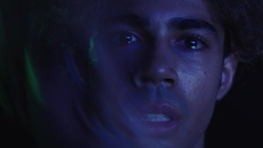 A young man sits in a dark room faint light reflecting on him revealing his face filled with sadness and tears in his eyes
