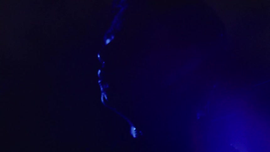 A mans face is faintly illuminated in deep blue light partially hidden in the darkness as he gets rained on