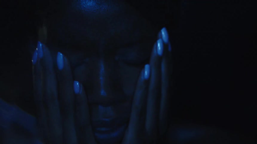 A woman in the dark under dim and flashing blue lights has her hands covering her face then removes them showing her sad expression