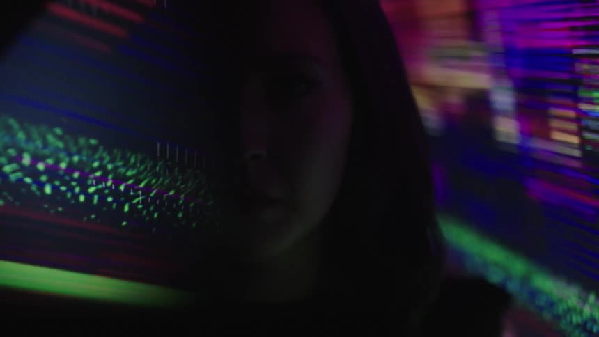 A kaleidoscope of flickering colors flows behind a woman with shoulderlength brown hair and half her face obscured in shadow and color staring straight towards the viewer