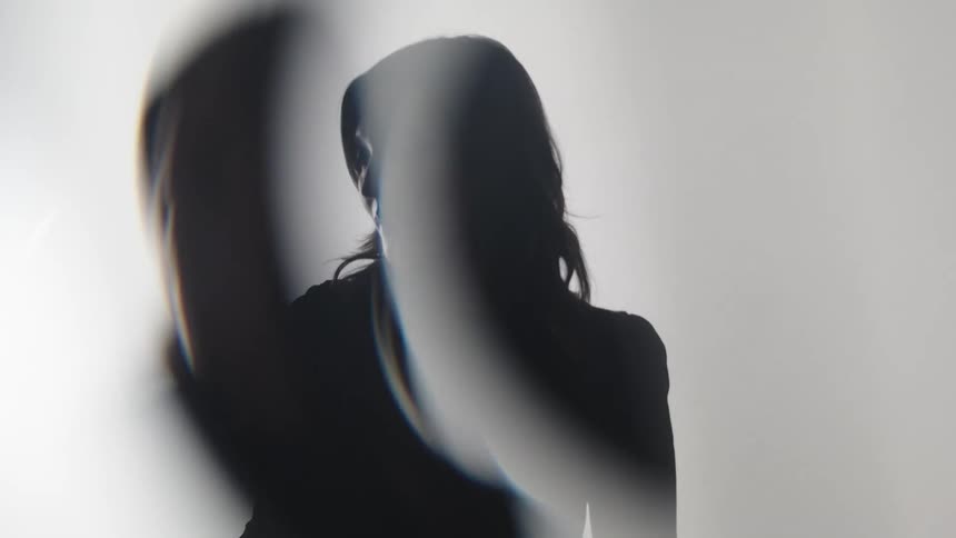 A blurry silhouette of a person with long dark hair is shown from the back turning their head to the left as a bright light shines from the left side of the frame