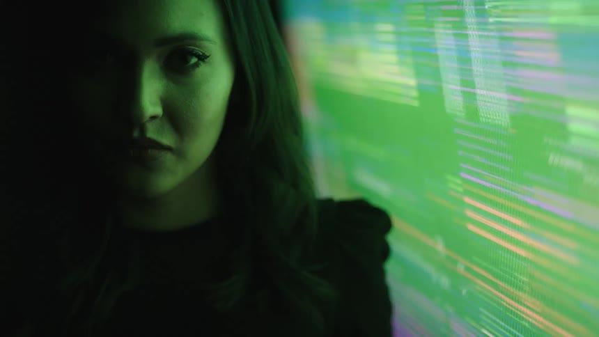 A womans face is seen in a close view with her long dark hair visible and her eyes looking directly into the camera as a colorful flashing screen illuminates her