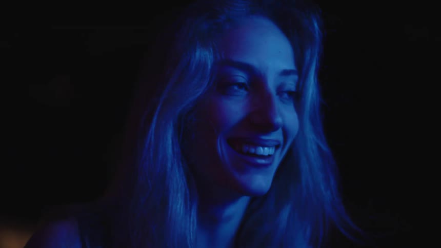 A young womans face bathed in a cool blue light smiling brightly in a lowlight dark setting
