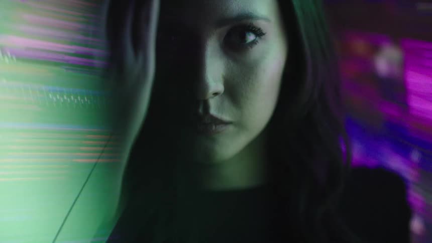 A closeup shot of a womans face is illuminated by flashing colorful lights that change rapidly as she stares intently at the camera