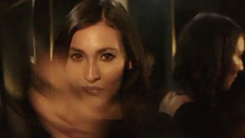 A beauty model with long hair is in a dark room as a flash of light briefly illuminates her face revealing her makeup