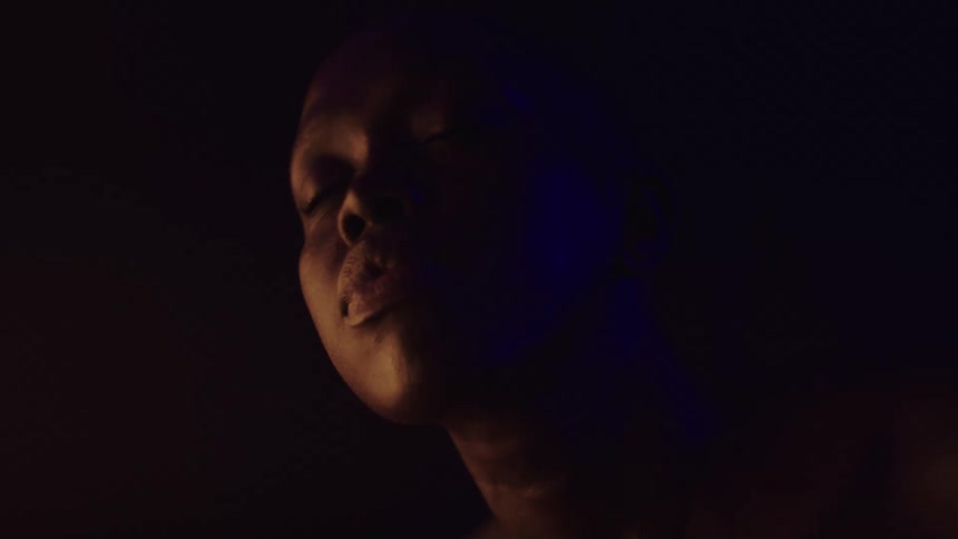 A close view of a woman with dark skin shows her turning her head to the left with an orange light on the left side of her face and a blue light on the right side