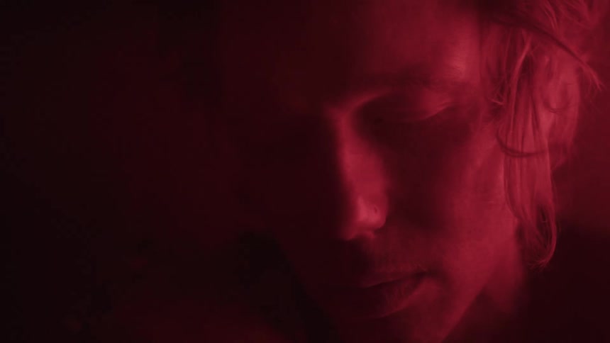 A young man wearing a wristwatch sits in a dark room illuminated by red light filled with sadness
