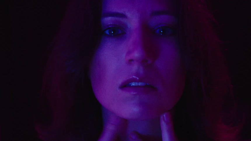 A womans face as she removes her hands revealing a serious expression under intense shifting purple and blue light