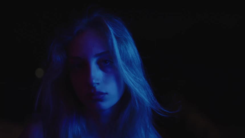 A close view shows a woman with long light colored hair and a serious expression looking directly into the camera while lit by a blue light
