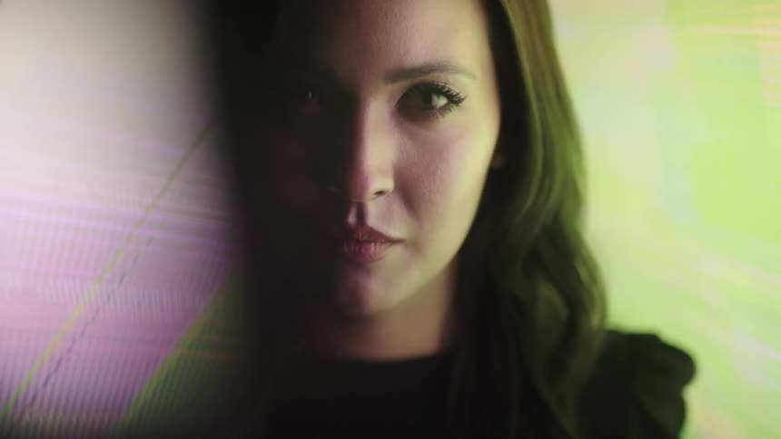 A young woman sits in a dark room deep in thought with a large monitor screen behind her