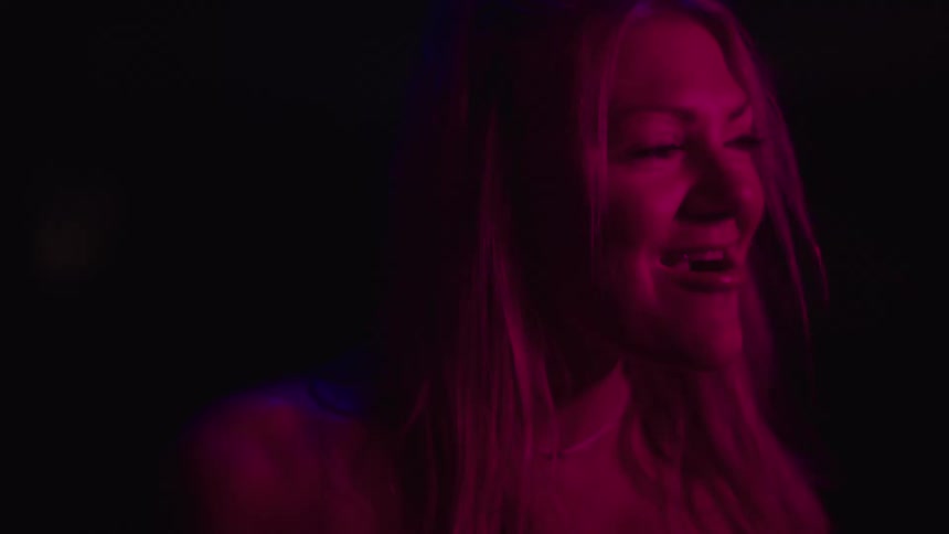A woman with bare shoulders is bathed in deep pink lighting smiling slightly as she looks off to the side