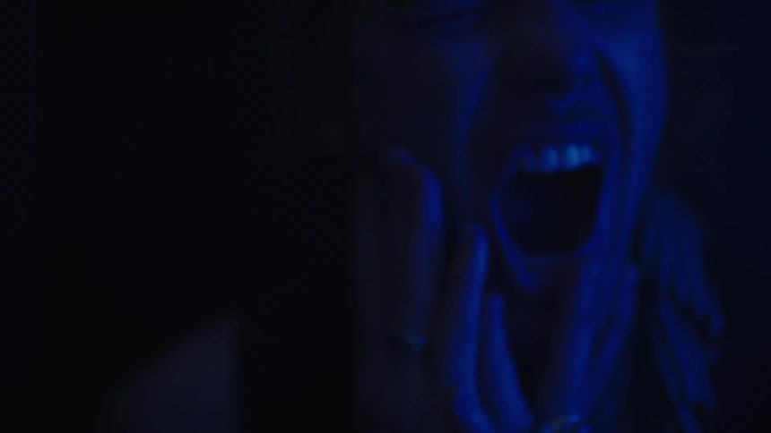 A person is yelling with their mouth wide open covering his face with his hands under a blue light