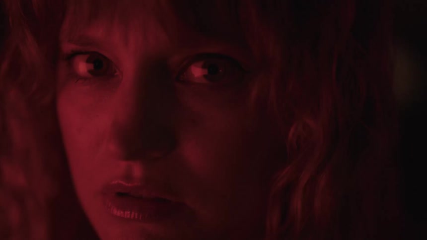 A closeup of a womans face illuminated by a deep red light shows her looking directly at the camera with a neutral expression