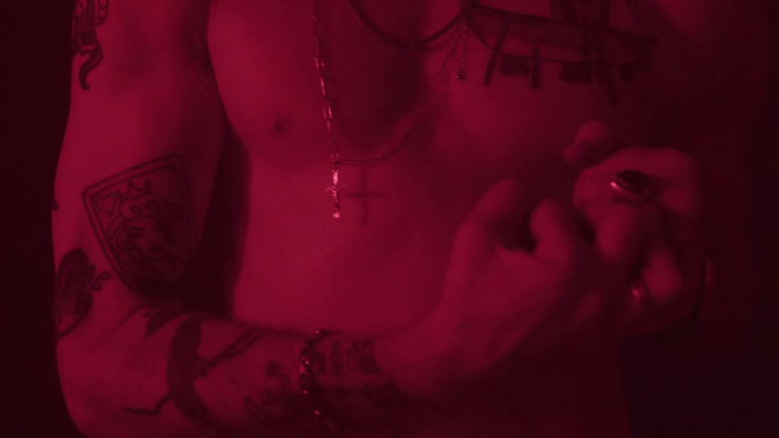 A close view of a mans bare torso shows several tattoos on his arms and chest as he clenches his fist while wearing multiple chains and a ring