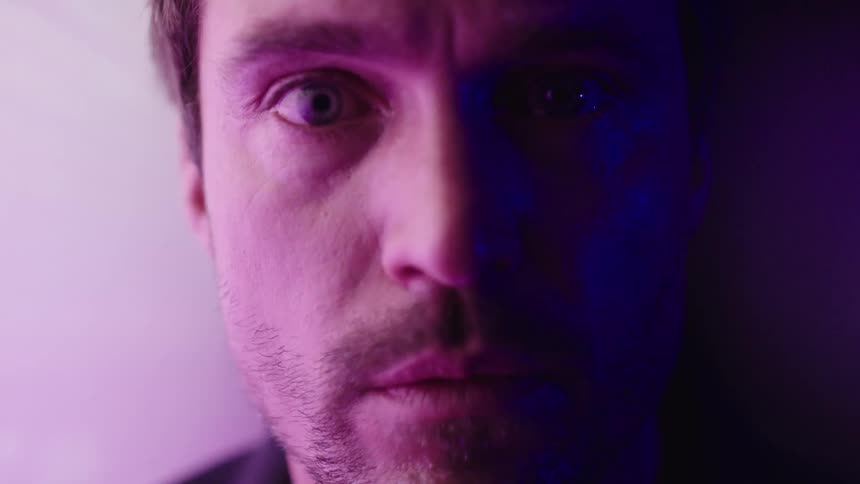 A young bearded man sits inside a room with a large screen to his left reflecting light that reveals his face