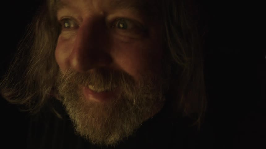 A closeup of a mans face with a beard and long hair smiling in a dark environment with light on his face