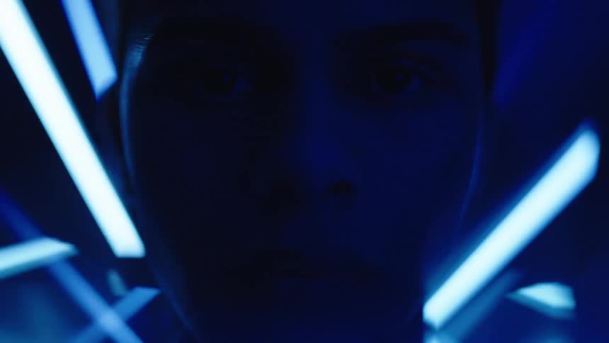 A womans face is seen in a close view with her eyes looking directly into the camera as a series of blue lights flash around her