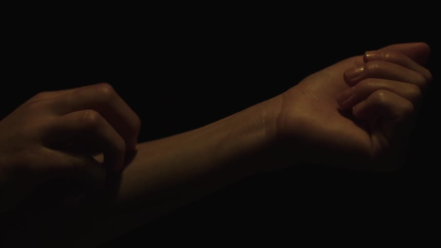A closeup of a persons hands is shown against a dark featureless background one hand is scratching the wrist of the other with a glimmer of light reflecting off a small ring on one finger