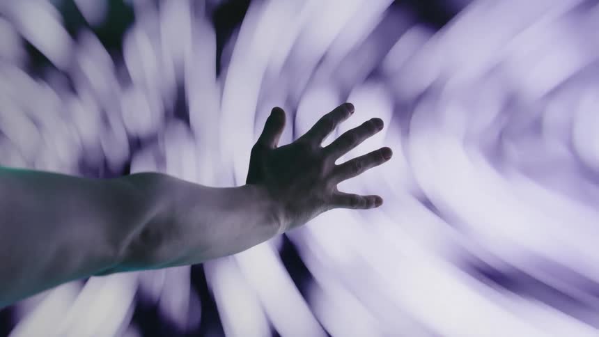 A very close view of a persons hand with their fingers spread out moving toward a blurry spinning purple and white lights in the background