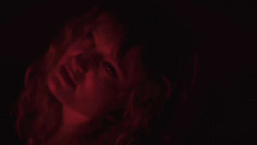 A young woman with long curly hair is bathed in red light she turns her head and looks into the camera smiling slightly