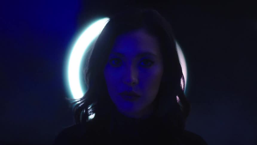 A young woman is shown in a closeup shot looking directly at the camera with a neutral expression she is in a dimly lit setting with a bright circular white light behind her head