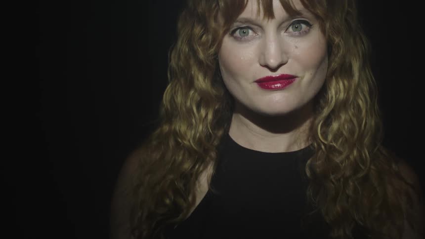 A closeup of a womans face with red lipstick and bangs as she looks directly at the camera with a subtle smile