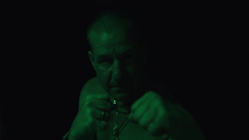 A man with short hair and a chain necklace boxes with his fists clenched while a green light illuminates his face and shoulders against a dark background