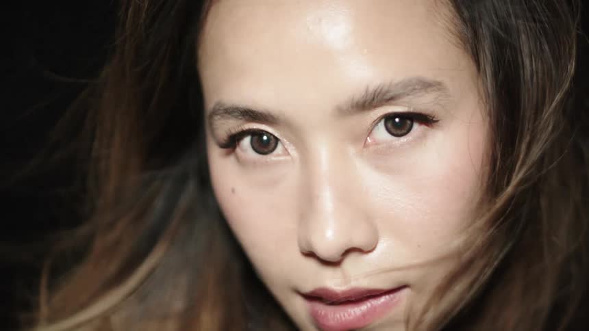 A young woman sits in a dark room as a bright flickering light briefly illuminates her face