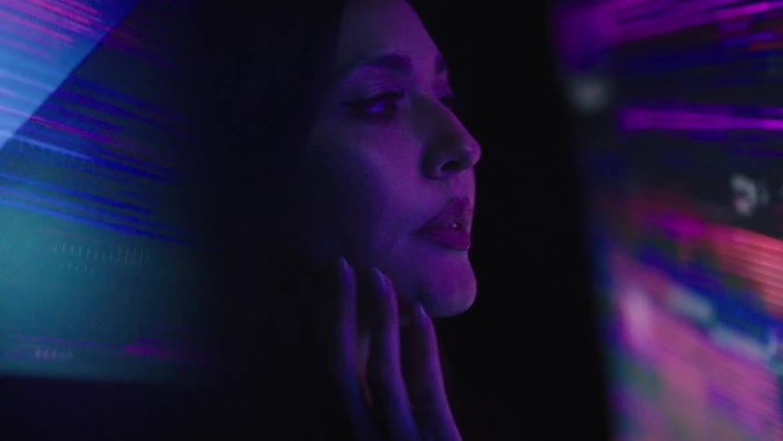 A woman with long dark hair is shown in a close view with her chin resting on her hand as a colorful flashing screen illuminates her face and she watches something off to her right