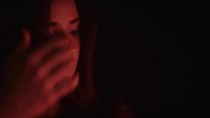 A woman lit by warm red light gazes downward with a pensive expression touching her hair