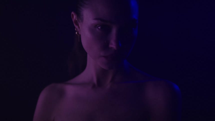 A young woman stands inside a dark room illuminated by dim blue light wearing earrings