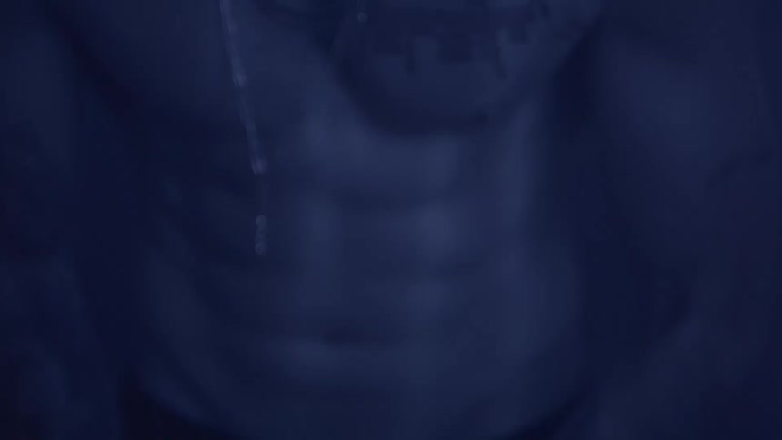 A shirtless man with defined abs and chest tattoos is illuminated by dim blue lighting