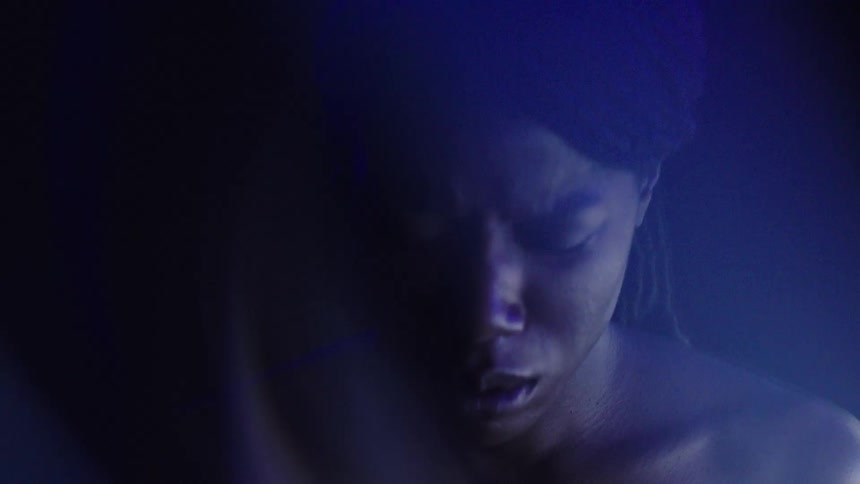 The video shows a person with a dark complexion and bare shoulders in a dimly lit space their face is partially visible and illuminated by a pulsing purple light