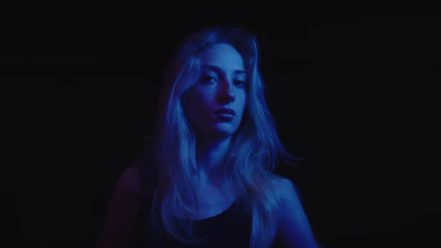 A young woman with long hair sits in a dark room wearing a black tank top deep in thought as the room is dimly illuminated by blue light