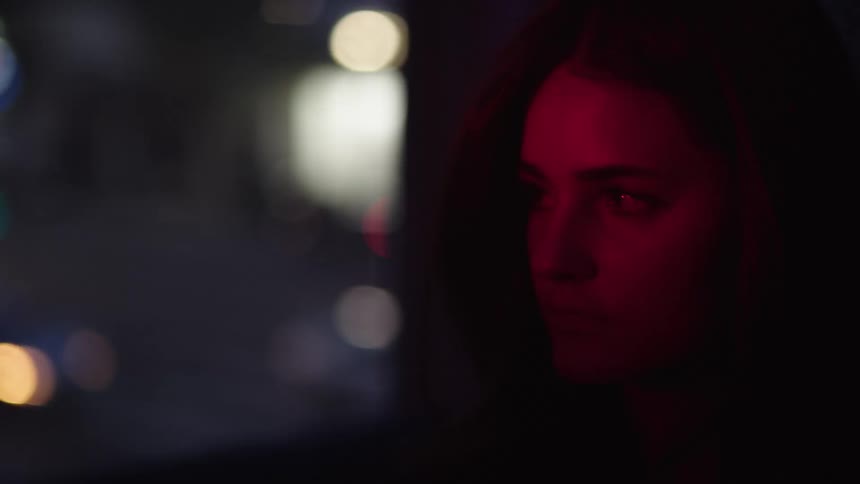 A closeup profile shot of a woman with long dark hair her face lit by a red light as she looks down and to the left with a serious expression with car passing in a blurry background