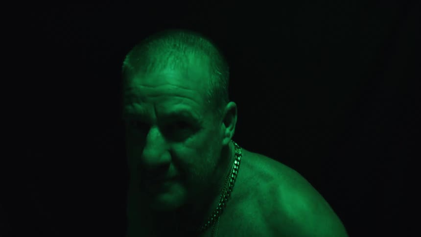 A middleaged man shirtless and wearing a silver necklace shadowboxes in a dark room illuminated by dim green light