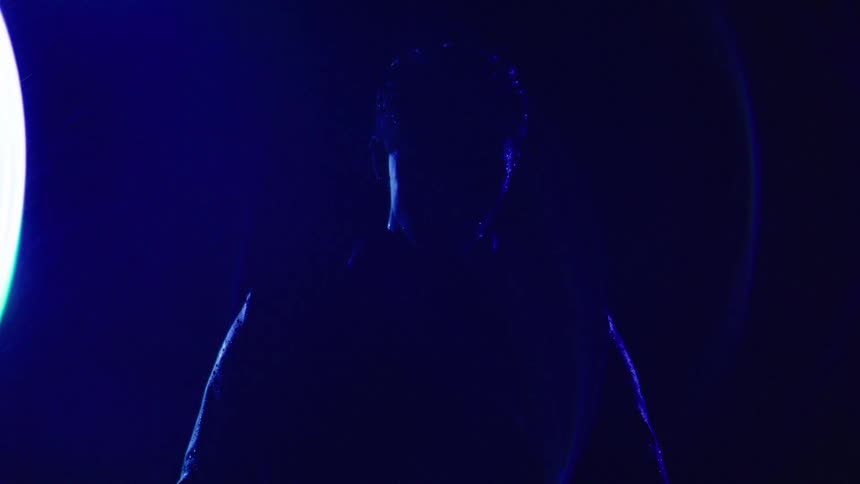 A man stands in deep shadow his outline faintly illuminated by intense blue light as it rains
