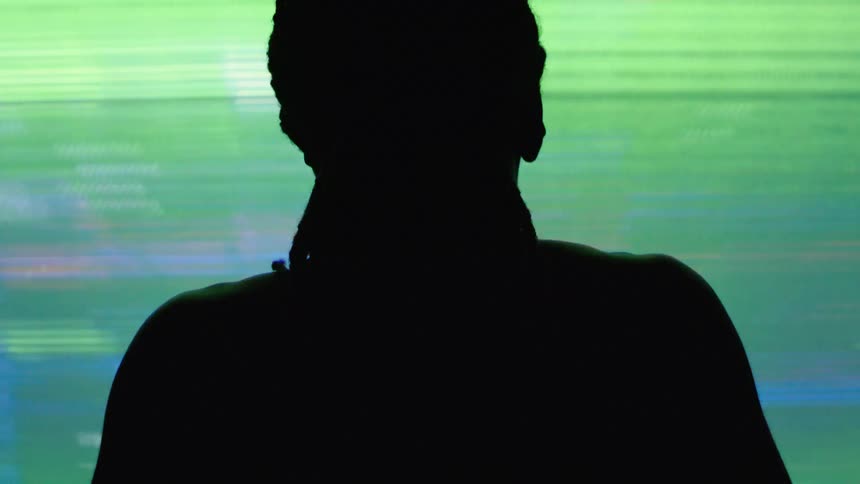 The video shows the silhouette of a person from behind with their head and shoulders visible the person is standing in front of a bright colorful and pixilated screen that illuminates the scene with pink and purple light
