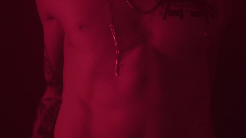 A shirtless man wearing a necklace with a cross pendant and tattoos on his chest and arm stands in a dark room illuminated by red light