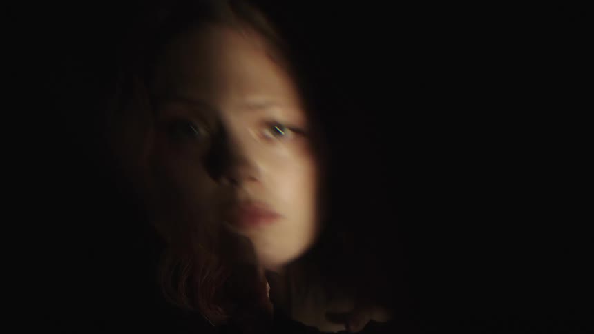 A dramatic closeup of a persons face is shown in a dark setting with only one side lit by a bright warm light the person looks directly at the camera with their hands clasped and then moving with some streaks of dirt or paint on their skin