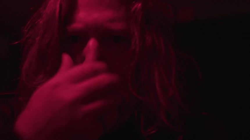 Longhaired man lit by intense red light stares at the camera with a serious expression