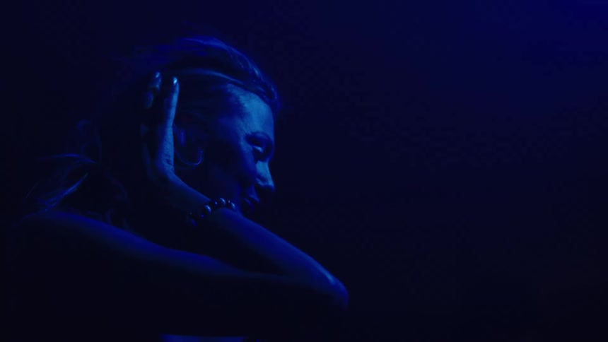 A woman bathed in deep blue light tilts her head back with her mouth open and hand on her chest as she dances