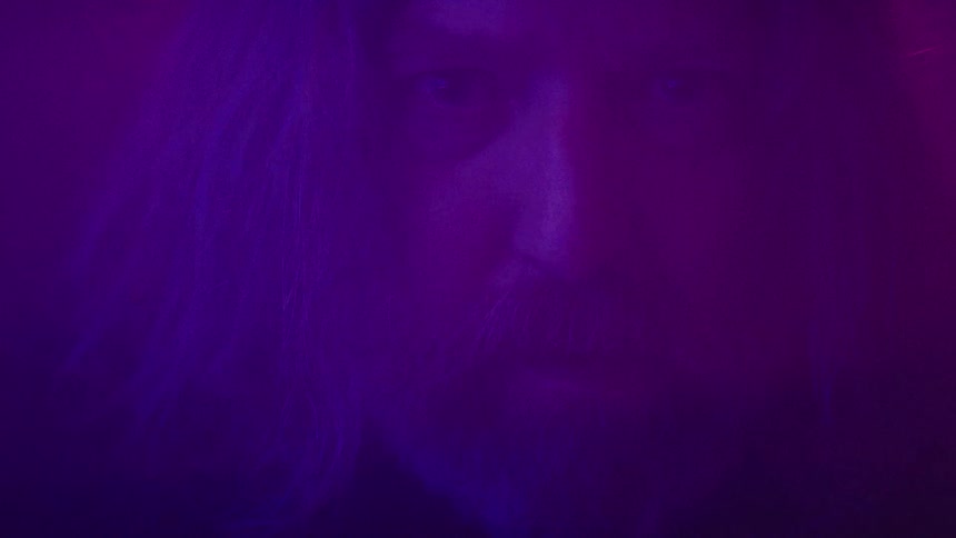 A man with long hair and a beard stands in a dark room illuminated by blue light looking furious