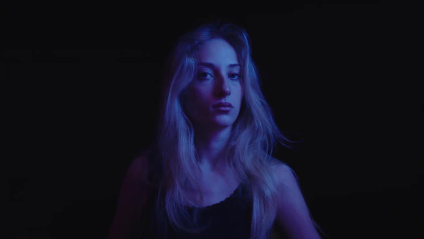 A woman with light colored hair and a dark shirt looks up to her right then turns her head to face the camera with a serious expression with a blue light illuminating her from the side