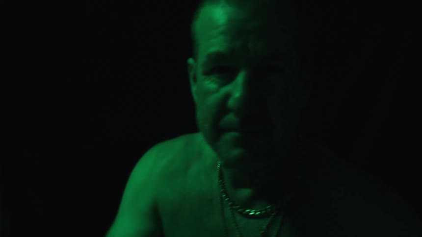 The video shows a man with a shaved head and a chain around his neck the scene is dark and the mans face and shoulders are illuminated by a green light